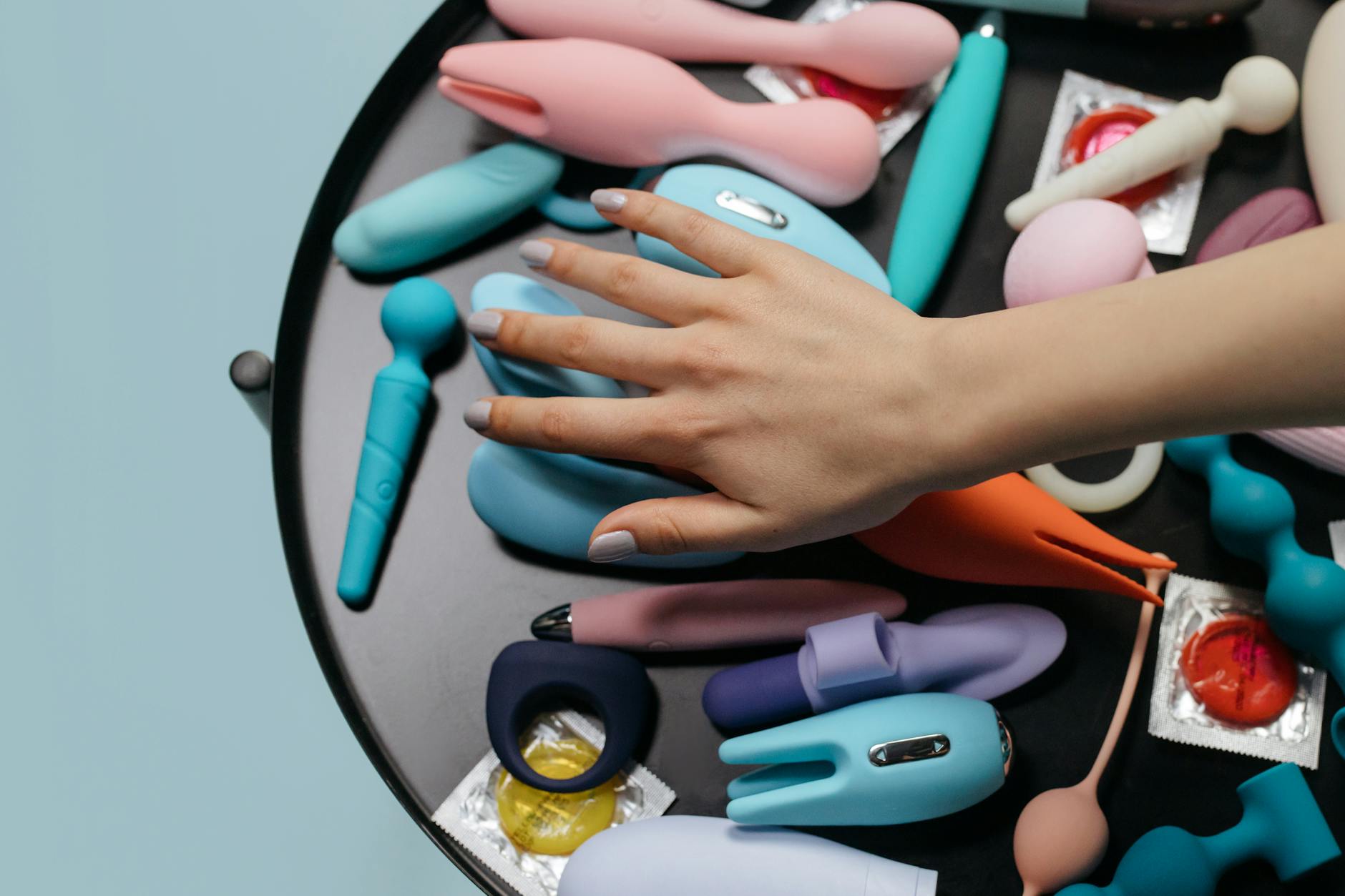 A hand selecting a clitoral vibrator from a collection of colorful adult toys on a table