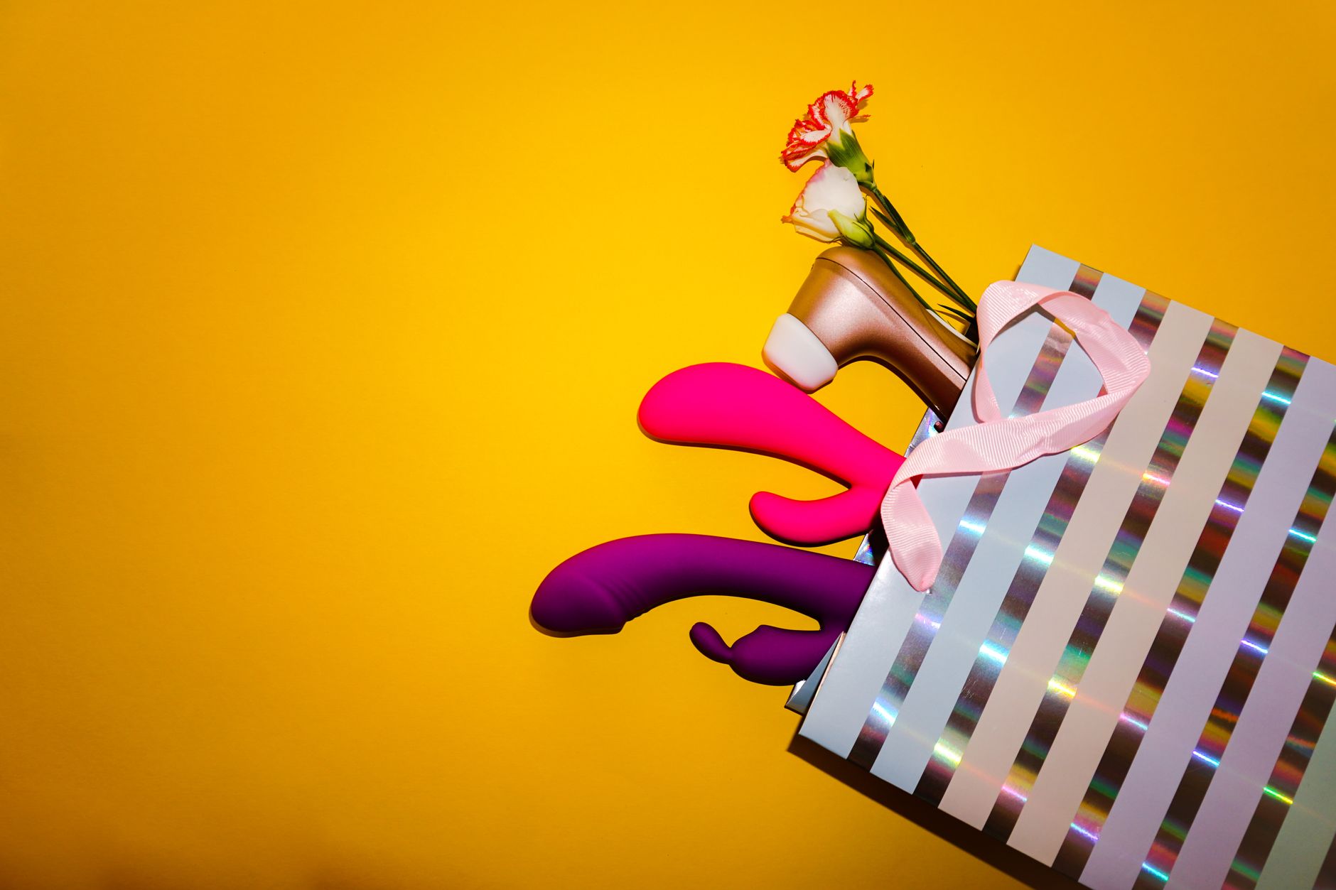 Colorful vibrators with flowers in a holographic gift bag set against a bold yellow background